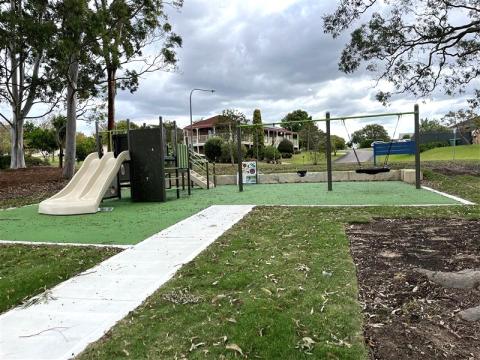 A photo of Applegum Park at Woongarrah with a play unit with a double slide, height chart, net bridge, climbing walls, sensory board, standard swing, nest swing and junior swing, full rubber softfall and pathway.
