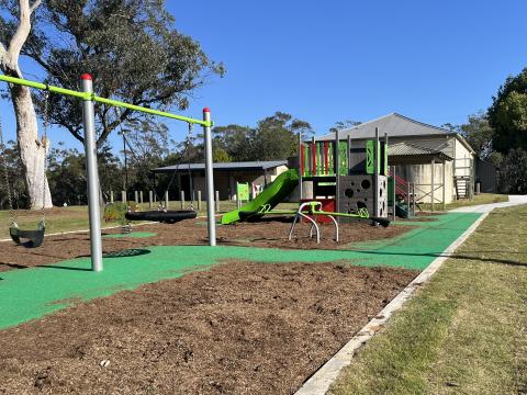A photo of a playspace in Pioneer Park, Kulnura with with three slides, a climbing net, standard swing, nest swing and junior swing, sensory panels, a seesaw, rocker, rubber and mulch softfall and pathway.