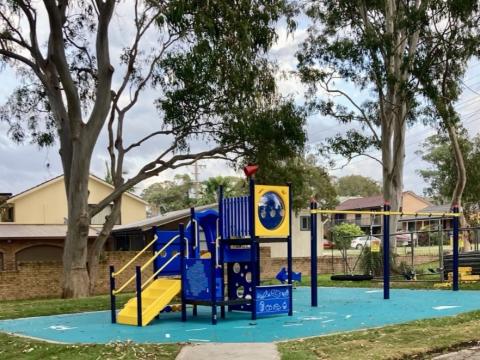 A photo of Lions Park in Gwandalan with a play unit with a slide, sensory play panels, a standard swing, nest swing and junior swing, a spring toy, rubber softfall and a pathway linking to an existing path.