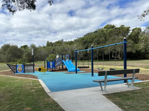 A photo of Norah Head playspace with a ship-themed play unit with a double slide climbing net, seesaw, rocker, standard swing, nest swing and junior swing, mulch softfall, rubber softfall and pathway.