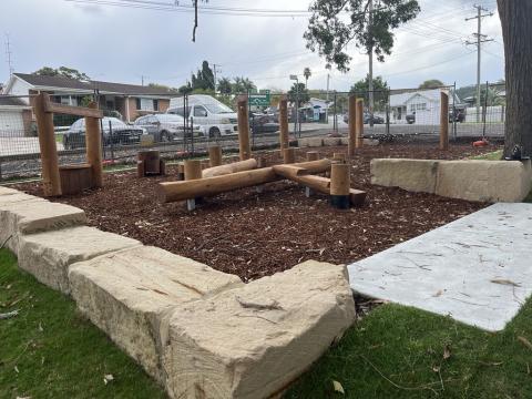 A photo of Wamberal Hall playspace with a nature play unit including tri net low climber, café shop front wooden drums, timber balance log combo and a rope balance course.