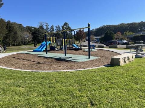 A photo of Woodbury Estate Park with a play unit with double slide and single slide, shop counter, climbing walls, bird rocker, standard swing, nest swing and junior swing, rubber and mulch softfall, sandstone seating and pathway.