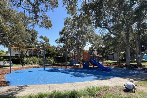 Blue softfall with colourful play equipment comprised of a swing set, slides and play unit. in the foreground there is a child crouched over playiing in the grass.