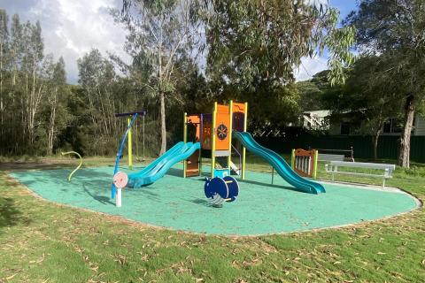 Colourful play equipment including slides, play unit and rockers on green softfall floor. Trees in background.